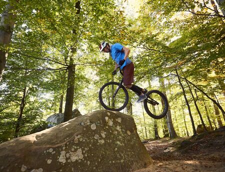 Professional cyclist riding on front wheel on trial bicycle. Sportsman biker balancing on the edge of big boulder in the forest on sunny day. Concept of extreme sport active lifestyleの写真素材