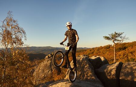 Athlete cyclist balancing on back wheel on trial bicycle. Sportsman rider looking to the camera, making acrobatic trick on the edge of big boulder on the top of mountain. Concept of extreme sportの写真素材