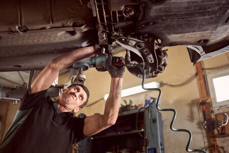 Repairman fixing car in garage. Experienced specialist car mechanic standing under lifted car during repair and maintenance process in repair station. Workshop for automobile checking up and repairingの写真素材