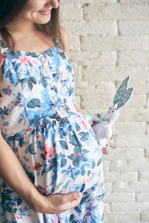 Side view of happy pregnant woman in dress standing, embracing big tummy and keeping lovely toy bunny near belly. Young cute future mother waiting for child. Concept of love and pregnancy.の写真素材
