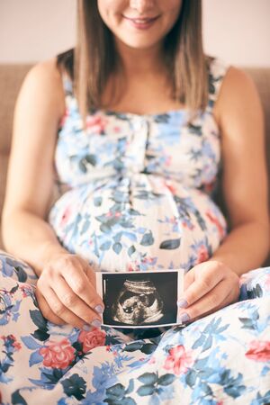 Smiling young woman sitting on sofa and keeping in hands ultrasound image of baby. Cheerful pregnant mother in dress waiting for infant. Concept of new life and expectation.の写真素材