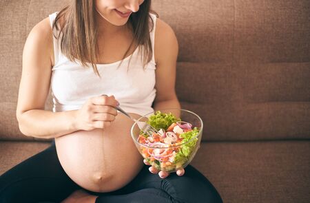 Front view of pregnant woman sitting on sofa at home and eating fresh salad. Happy mother eating vegetables and caring about health and baby. Concept of expectation and health.の写真素材