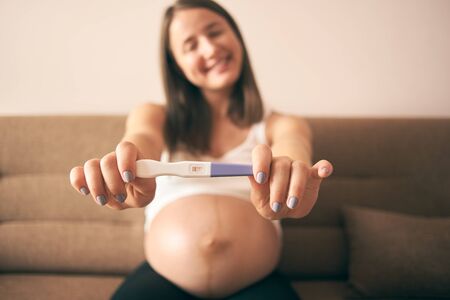 Selective focus of pregnancy test in hands of smiling woman in white shirt. Happy future mother enjoying motherhood and waiting for little baby. Concept of expectation and happiness.の写真素材