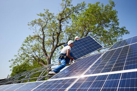 Two workers in protective helmet on tall steel platform mounting heavy solar photo voltaic panel on green tree and blue sky background. Exterior solar panel system installation, dangerous job concept.の写真素材
