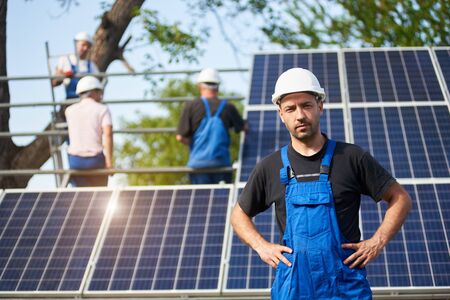 Portrait of smiling successful engineer technician standing in front of unfinished high exterior solar panel photo voltaic system blue shiny surface, with team of workers on high platform.の写真素材