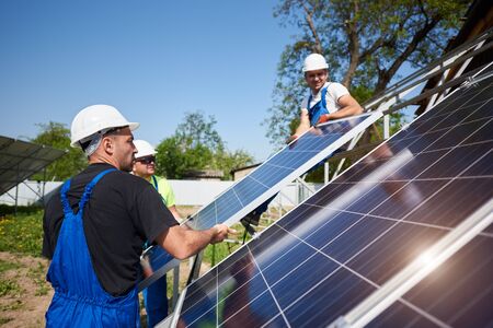 Team of three professional workers installing heavy solar photo voltaic panels on high steel platform. Exterior solar system installation, alternative renewable green energy generation concept.の写真素材