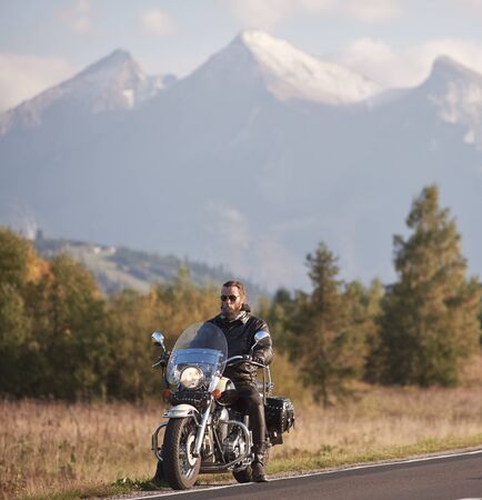 Bearded biker in black leather jacket and sunglasses sitting on cruiser bike on country roadside on blurred background of green rural landscape, distant white sunny mountain peaksの写真素材