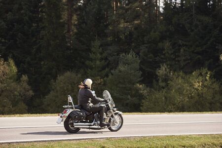 Side view of long-haired motorcyclist in helmet and black leather clothing riding cruiser motorbike along asphalt path, on sunny summer day on background of tall trees.の写真素材