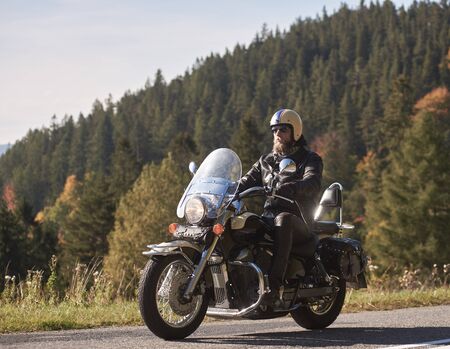 Handsome bearded biker in black leather jacket and sunglasses sitting on cruiser motorcycle on country roadside, on blurred background of foggy green hills covered with dense spruce forest.の写真素材