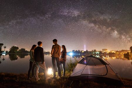 Night camping on lake shore near campfire. Two couple having a rest near tourist tent together. Friends enjoying beautiful view of night sky full of stars and Milky way, luminous town on background.の写真素材