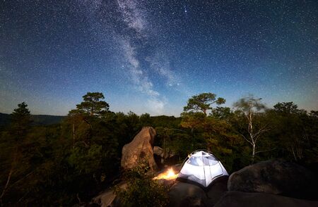 Camping at summer night on rocky mountain. White tourist tent and bonfire under magical night sky full of stars and Milky way. On the background beautiful starry sky, big boulders and treesの写真素材