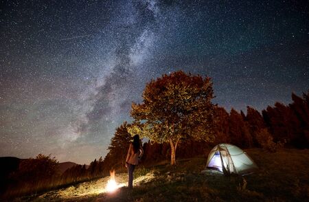 Back view of woman tourist resting at campfire beside camping and illuminated tourist tent, enjoying view of night sky full of stars and Milky way. On background starry sky, mountains and big treeの写真素材