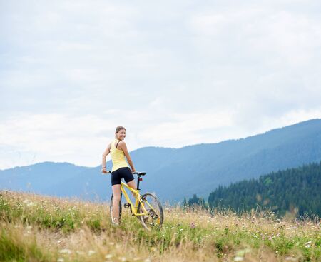 Back view of attractive happy woman cyclist riding on yellow mountain bicycle on a grassy hill, enjoying summer day in the mountains. Outdoor sport activity, lifestyle conceptの写真素材