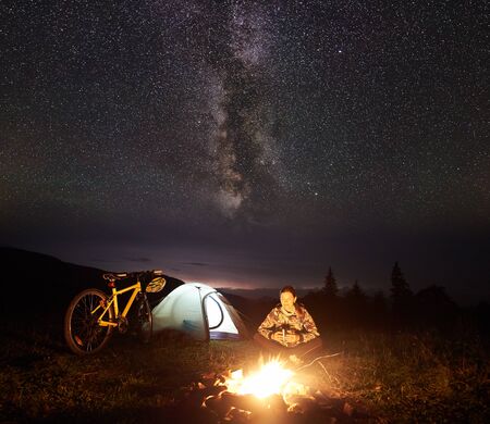Young woman cyclist resting at night camping near burning campfire, illuminated tourist tent, mountain bike under incredible beautiful evening sky full of stars and Milky way. Tourism conceptの写真素材