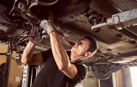 Repairman fixing car in garage. Experienced specialist car mechanic standing under lifted car during repair and maintenance process in repair station. Workshop for automobile checking up and repairingの写真素材