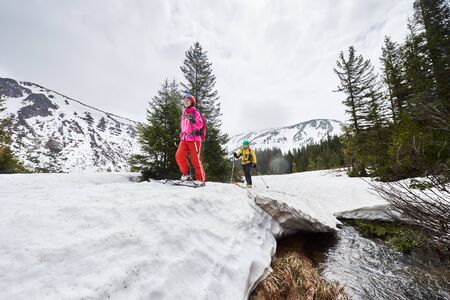 Ski touring and off-piste concept. Small group of people sliding on ski on snow-covered bridge and flowing stream under picturesque snow-covered mountains and coniferous forest on backgroundの写真素材