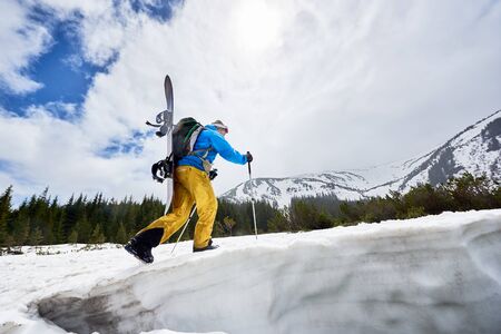 Side of snowboarder with hidden face carrying snowboard on his back and ascending snowy slope up to mountain peak. Adventure in mountains. Clouds on blue sky on background. Snapshot from low angleの写真素材