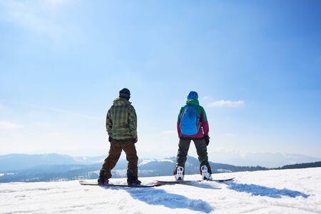 Back view of two tourists snowboarders in sportive equipment standing on snowboards on copy space background of blue sky and woody mountains on sunny winter day. Extreme winter sports concept.の写真素材