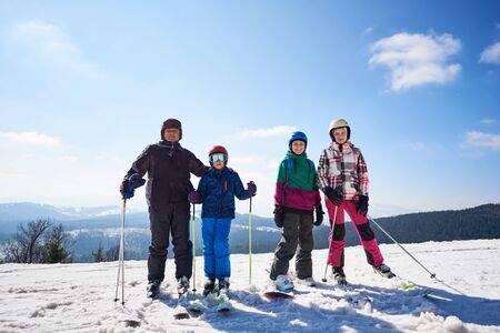 Photo of sportive happy family outdoors on sunny winter day. Group of smiling man and three girls on skis and snowboard standing in deep snow on copy space background of blue sky and woody mountains.の写真素材