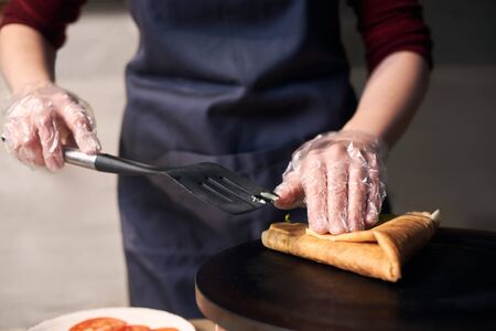Rolled up big pancake like triangle made on hot round professional cooktop by chef in gloves. Cropped hands with black spatula cooking big crepe stuffed with vegetables. Focus view. Blurred backgroundの写真素材
