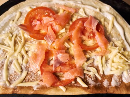 Cutted slices of raw salmon fish and tomato, grated cheese on round thin and baked dough base greased with white sauce. Black round tray on background. Italian cuisine concept. Focus detailed top viewの写真素材