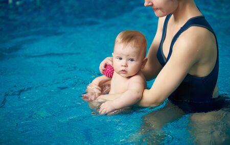 Cropped view of mother and son in swimming pool. Sweet baby with toy in hand, sitting in his mothers arms during sport activity, exercising for child development in water and looking on cameraの写真素材