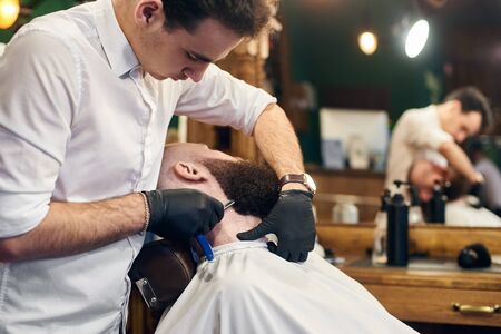 Man client reclining in chair for shaving by barber hands in gloves with sharp blade on bearded face. Trust, skillful hands, servicing, barbershop concept. Blurred backgroundの写真素材
