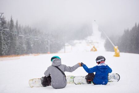 Back view of sportsmen couple sitting on snow before freeriding from ski run at ski resort. Man and woman doing fist bump. Snowfall in mountains. Friendship, support, decision, sport, extreme conceptの写真素材