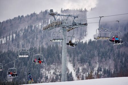 Bukovel, Ukraine - December 09, 2018: skiers and snowboarders sitting carrying high up on cable chairlift in one direction over snow-covered slopes in Carpathians mountains in foggy gray dayのeditorial素材