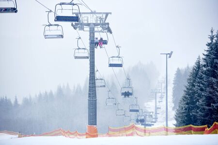 Chairlift using for carrying passengers skiers and snowboarders up and down in mountains. Fir and pine trees around. Foggy visibility in mountains. Organization of rest and skiing at ski resort.の写真素材