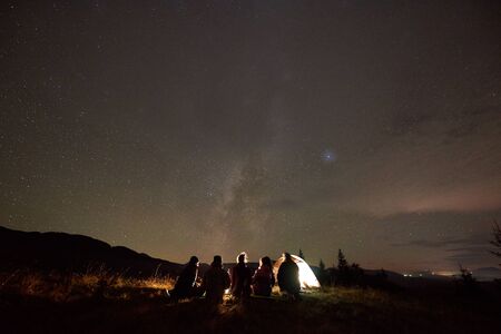 Night camping under stars, tourism and active lifestyle. Back view of five people sitting on grassy valley at tourist tent and burning campfire on copy space background of dark starry sky.の写真素材