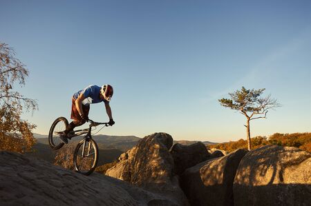Young cyclist standing on front wheel on trial bicycle. Professional sportsman rider balancing on the edge of big boulder on the top of mountain at sunset. Concept of extreme sport active lifestyleの写真素材