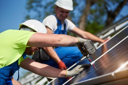 Close-up of two professional technicians connecting solar photo voltaic panel to metal platform on blue sky background. Stand-alone solar system installation, efficiency and professionalism concept.の写真素材