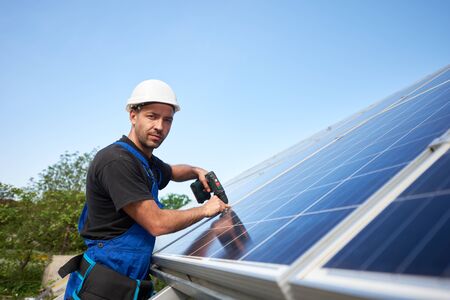 Technician installing solar panel to metal platform using electrical screwdriver on blue sky copy space background. Stand-alone solar system installation, efficiency and professionalism concept.の写真素材