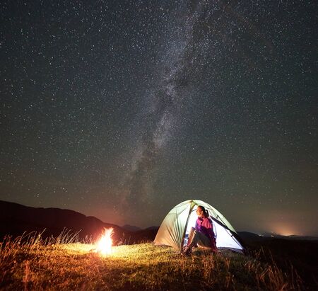 Female hiker resting at summer night camping in the mountains beside bonfire. Happy woman traveller sitting in illuminated tourist tent, enjoying view of night sky full of stars and Milky way.の写真素材