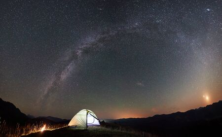 Panoramic view of tourist camping at night in the mountains. Illuminated tent and campfire under beautiful night sky full of stars and Milky wayの写真素材