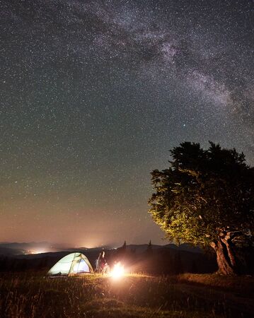 Female hiker relaxing at summer night camping in the mountains beside bonfire, illuminated tourist tent and big tree. Woman sitting on chair, enjoying view of night sky full of stars and Milky way.の写真素材