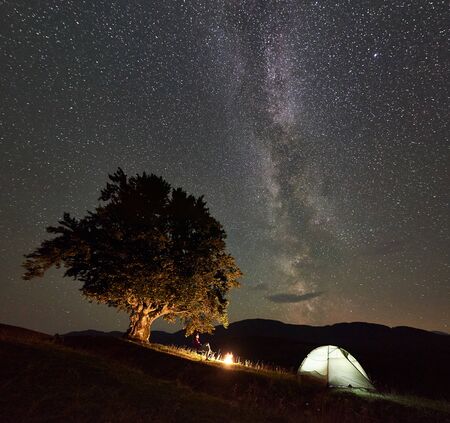 Female hiker relaxing at summer night camping in the mountains beside bonfire and glowing tourist tent. Traveller sitting on chair under big tree and beautiful night sky full of stars and Milky way.の写真素材