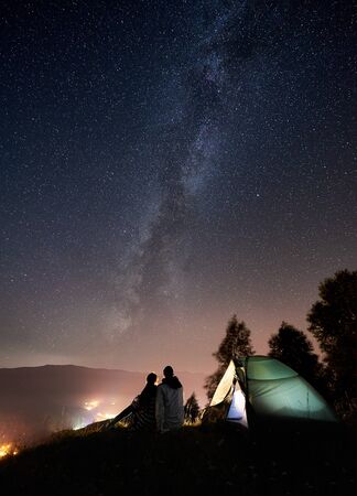 Back view of young couple tourists having a rest at bonfire near illuminated tent under amazing night sky full of stars and Milky way. On background beautiful starry sky, mountains and luminous cityの写真素材