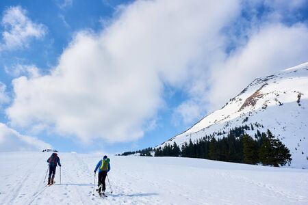 Back view of two hikers, skiers with backpacks trekking on skis up snowy hill on copy space background of bright blue sky and beautiful mountains on frosty sunny day. Winter active vacations concept.の写真素材