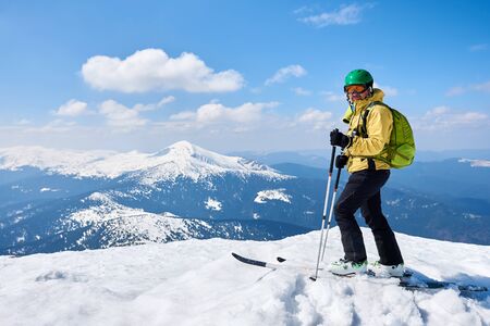 Sportsman skier in helmet and goggles with backpack standing on skis holding ski poles in deep white snow on copy space background of bright blue sky and highland landscape. Winter skiing concept.の写真素材