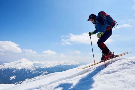 Sportsman skier in skiing equipment with backpack jumping in air down steep snowy mountain slope on copy space background of blue sky and highland landscape. Winter sports, courage and speed concept.の写真素材