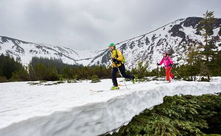 Couple of cross country skiers man and woman with backpacks walking and exercising on ski in mountains near winter coniferous forest. Snow-covered mountain in full altitude on backgroundの写真素材