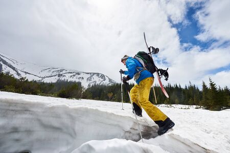 Side view of male in winter mountain touring walking up with ski poles helping and carrying backpack and snowboard on his back. Ski touring and mountaineering promotion. Landscape view with copy spaceの写真素材