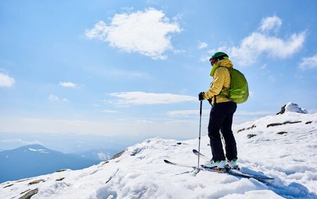 Back view of sportsman skier in helmet and goggles with backpack standing on skis holding ski poles in deep white snow, on copy space background of bright blue sky enjoying beautiful mountain view.の写真素材