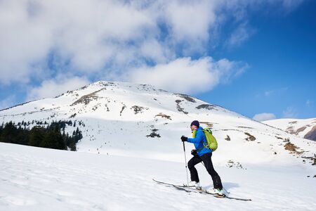Male hiker, tourist in colorful clothing with backpack trekking on skis up snowy hill on background of bright blue sky and beautiful mountain on frosty sunny day. Winter active vacations concept.の写真素材