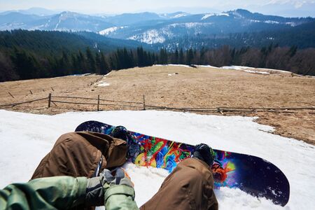 Point of view shot of snowboarder legs and bright colorful snowboard in snow, view of winter landscape. Mountain clearing among tall spruce trees, snowy peaks in background. Sports and pov concept.の写真素材