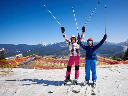 Two skiers, boy and girl in warm clothing and goggles on skis with raised poles in deep snow on background of winter ski resort, bright blue sky and beautiful mountains. Sports and recreation concept.の写真素材