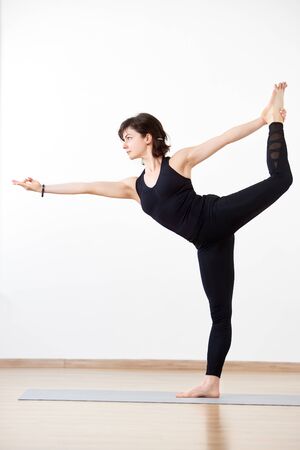 Flexible woman, yogi performing stance in popular Lord of dance pose. Female athlete in black on white background. Concept of attraction to perfection, body and mind unity. Vertical side portraitの写真素材