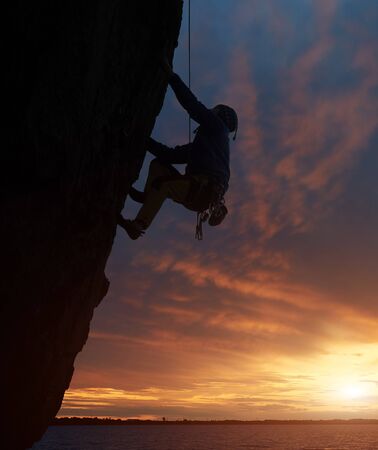 Silhouette of athletic person in safety harness hanging on edge of challenging cliff. Climber on rope rock climbing on cliff over water at sunset. Concept of mountaineering, adventure and challengeの写真素材